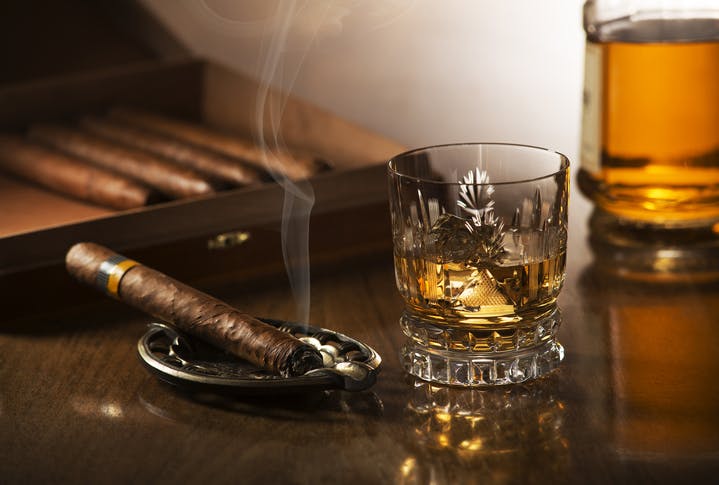 Image shows a smoking cigar on an ash tray beside a crystal glass containing whisky