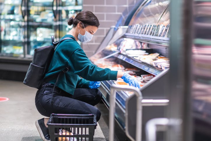 Image shows woman in surgical mask and gloves squatting in front of supermarket food shelves