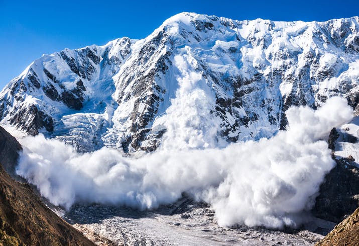 Image shows a white-out avalanche coming off a sunlit mountain against a blue sky