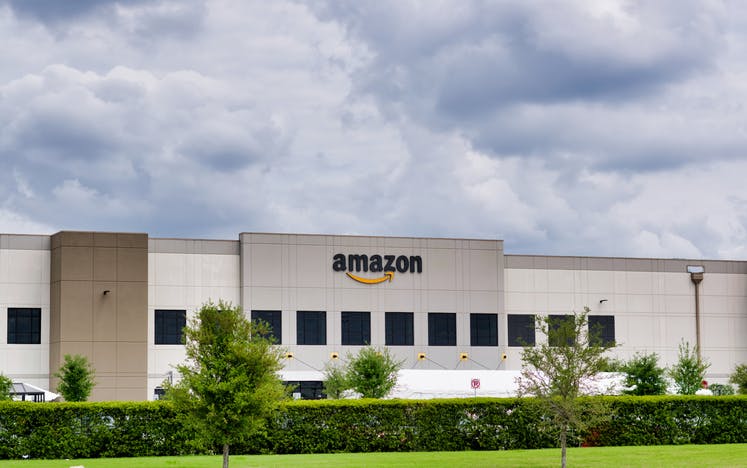 Image shows low-rise white Amazon building with greenery in the foreground against a light cloudy sky