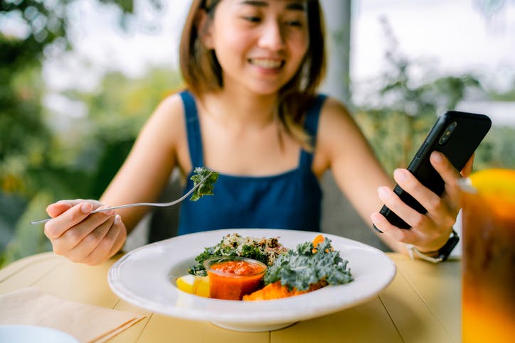 Image shows a young woman smiling and using her smartphone while eating a colourful plate of fruit and vegetables