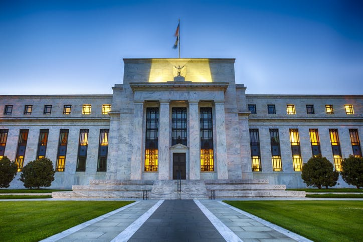 Image shows the front of the Federal Reserve buiding at dusk with lights showing through the windows