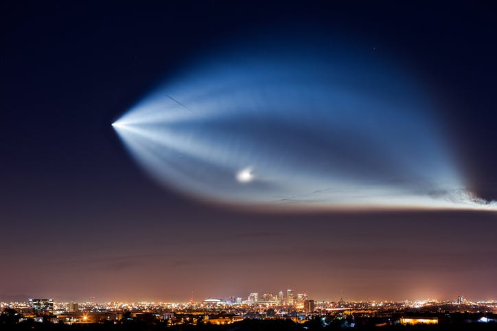 SpaceX Falcon 9 rocket launch showing billowing vapor trail in night sky over cityscape